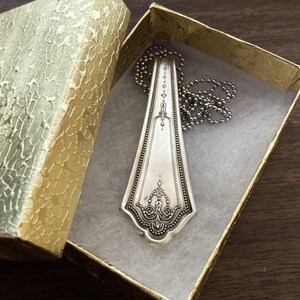 May include: A silver spoon necklace with an ornate design, displayed in a gold box with a white interior.