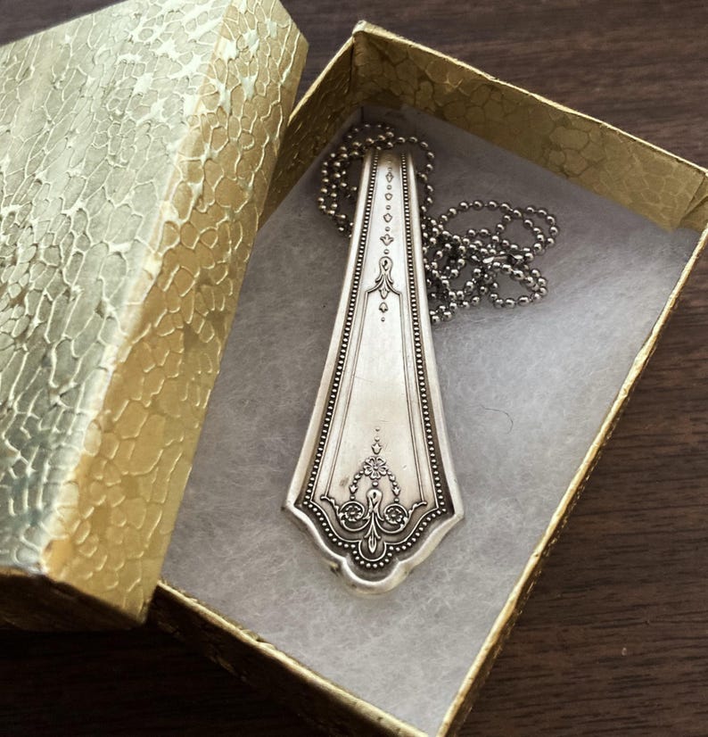 May include: A silver spoon necklace with an ornate design, displayed in a gold box with a white interior.