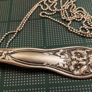 May include: A silver spoon necklace with a floral design. The spoon is attached to a silver chain.