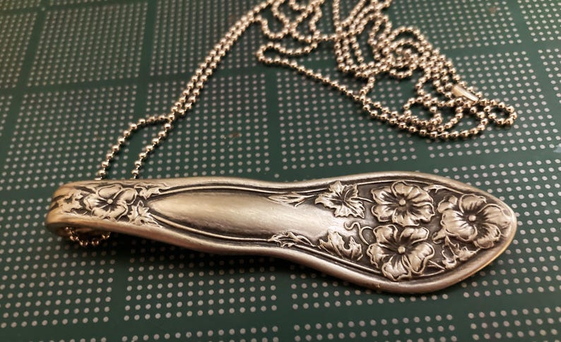 May include: A silver spoon necklace with a floral design. The spoon is attached to a silver chain.