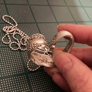 May include: A silver spoon bent into a heart shape with a chain attached. The spoon has a floral design.