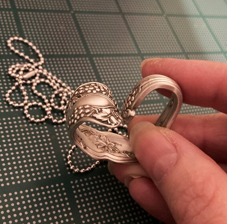 May include: A silver spoon bent into a heart shape with a chain attached. The spoon has a floral design.