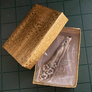 May include: A silver spoon necklace with a floral design, displayed in a gold box with a white lining.