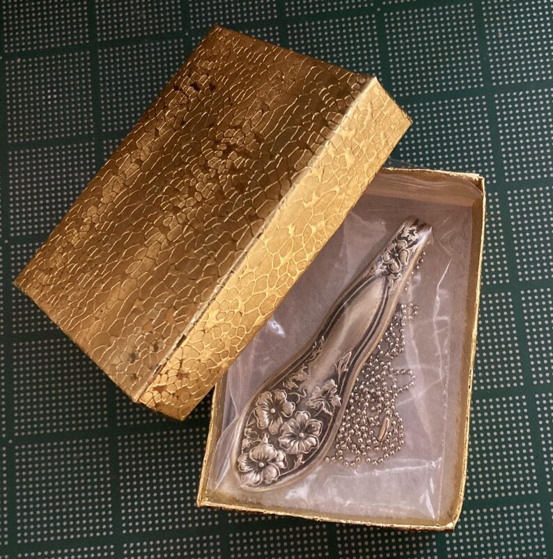 May include: A silver spoon necklace with a floral design, displayed in a gold box with a white lining.