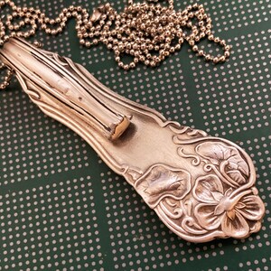 May include: A silver necklace with a pendant made from a vintage silverware piece. The pendant features a floral design with a delicate, intricate pattern. The chain is a simple ball chain.