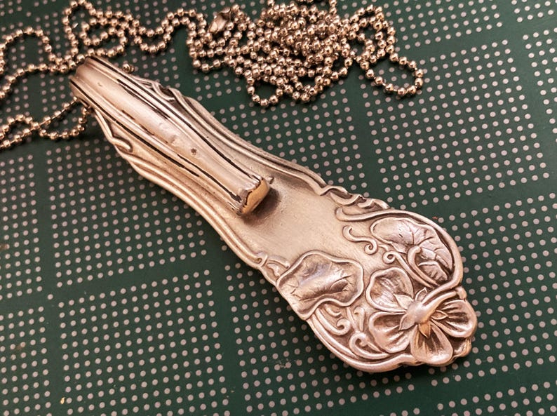 May include: A silver necklace with a pendant made from a vintage silverware piece. The pendant features a floral design with a delicate, intricate pattern. The chain is a simple ball chain.