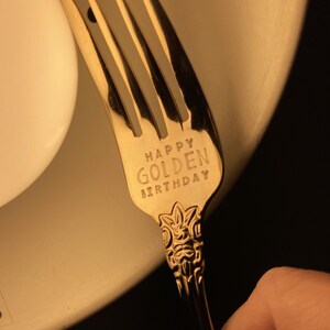 May include: A gold-colored fork with the words "HAPPY GOLDEN BIRTHDAY" engraved on the handle. The fork has a decorative design on the handle and is set against a soft, warm background.