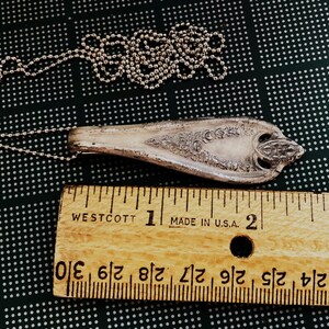 May include: A silver spoon necklace with an ornate design. The spoon is about 2 inches long and is attached to a silver chain. The chain is about 18 inches long. The spoon and chain are lying on a black and white patterned surface.