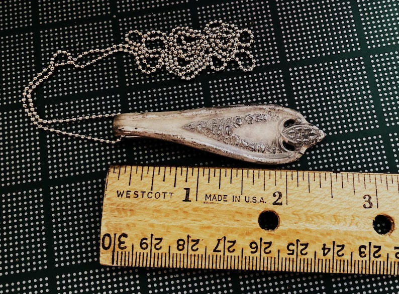 May include: A silver spoon necklace with an ornate design. The spoon is about 2 inches long and is attached to a silver chain. The chain is about 18 inches long. The spoon and chain are lying on a black and white patterned surface.