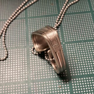 May include: A silver spoon necklace with a decorative design on the back of the spoon. The necklace is made with a silver ball chain.