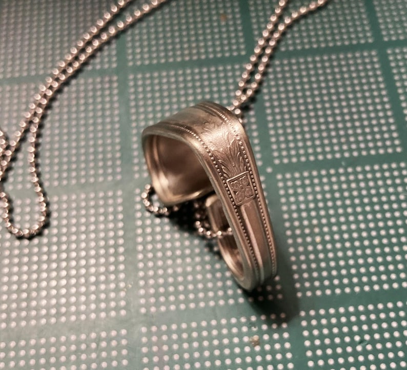 May include: A silver spoon necklace with a decorative design on the back of the spoon. The necklace is made with a silver ball chain.