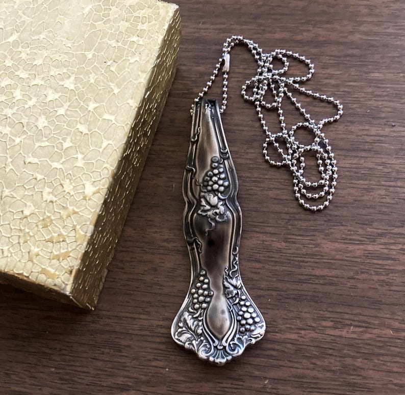 May include: A silver necklace with a pendant made from a vintage silverware piece featuring a grapevine design. The pendant is attached to a silver ball chain.