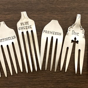 May include: Seven silver forks with different cheese names engraved on them. The names are Asiago, Mozzarella, Blue Cheese, Parmesan, Brie, and Pepper Jack.