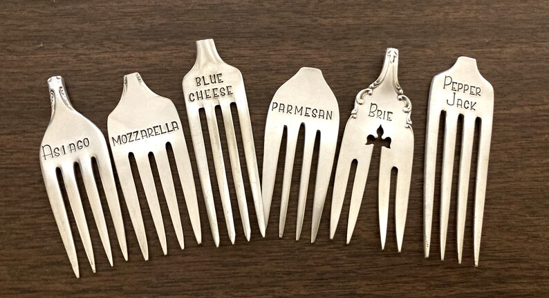 May include: Seven silver forks with different cheese names engraved on them. The names are Asiago, Mozzarella, Blue Cheese, Parmesan, Brie, and Pepper Jack.