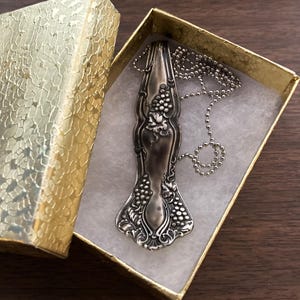 May include: A silver spoon necklace with a grape design. The necklace is in a small, gold gift box.