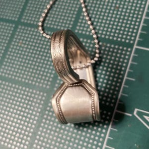 May include: A silver spoon necklace with a decorative design on the handle. The spoon is bent into a heart shape and is attached to a silver chain.