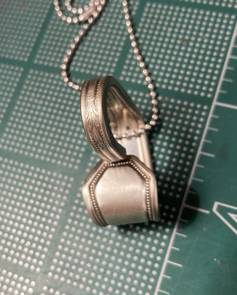 May include: A silver spoon necklace with a decorative design on the handle. The spoon is bent into a heart shape and is attached to a silver chain.