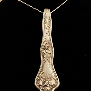 May include: A silver necklace with a pendant made from a vintage spoon. The spoon is decorated with floral designs and has a long, slender shape.