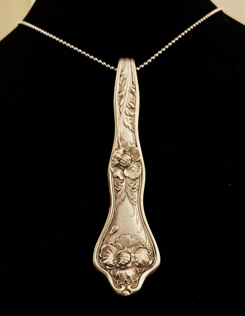 May include: A silver necklace with a pendant made from a vintage spoon. The spoon is decorated with floral designs and has a long, slender shape.