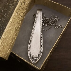May include: A silver spoon necklace with a decorative design and a silver chain. The necklace is in a gold box with a textured pattern.