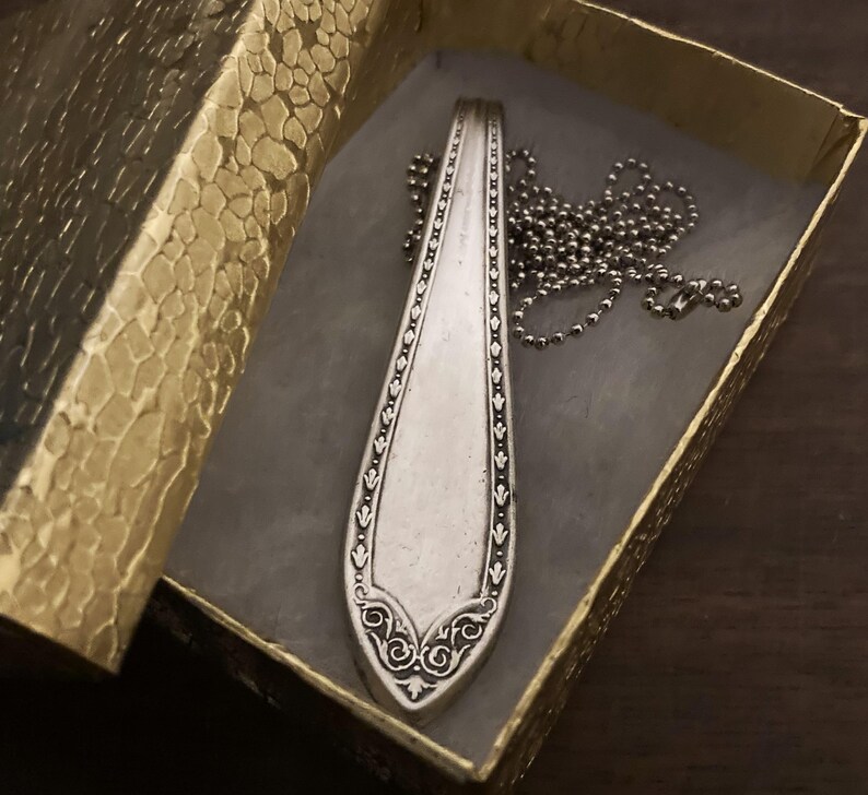 May include: A silver spoon necklace with a decorative design and a silver chain. The necklace is in a gold box with a textured pattern.