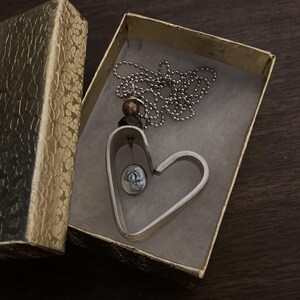 May include: A silver heart-shaped pendant necklace with a small, round, green and silver bead hanging inside the heart. The necklace is in a gold gift box.