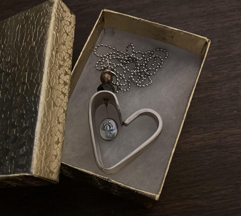 May include: A silver heart-shaped pendant necklace with a small, round, green and silver bead hanging inside the heart. The necklace is in a gold gift box.