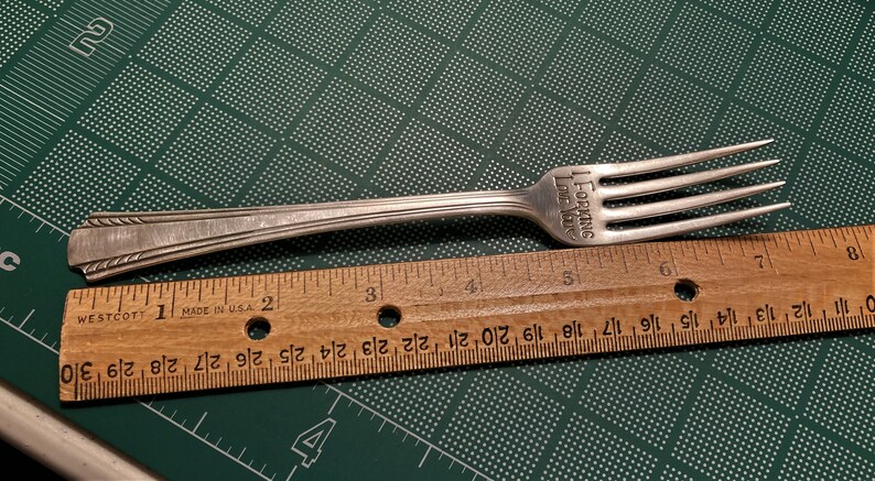 Op de afbeelding: Een zilveren vork met de inscriptie "I FORKING LOVE YOU" op het handvat. De vork is 16,5 cm lang en wordt gemeten tegen een houten liniaal.