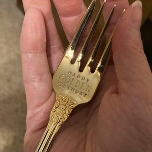May include: A gold-colored fork with the words "HAPPY GOLDEN BIRTHDAY" engraved on the handle. The fork has a decorative floral design on the handle and four tines.