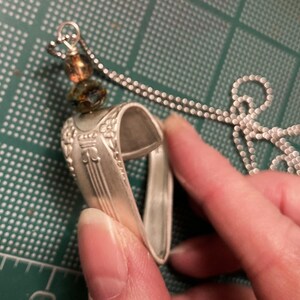 May include: A silver heart-shaped pendant necklace made from a repurposed spoon with a brown and green glass bead charm.