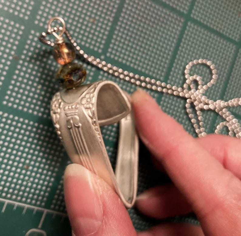 May include: A silver heart-shaped pendant necklace made from a repurposed spoon with a brown and green glass bead charm.