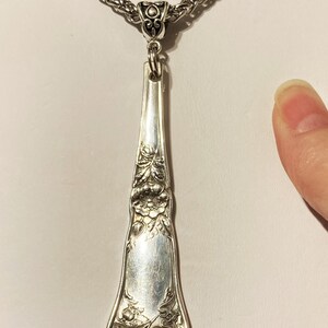 May include: A silver necklace with a long, ornate pendant made from a vintage silverware piece. The pendant features a floral design and is attached to a silver chain.