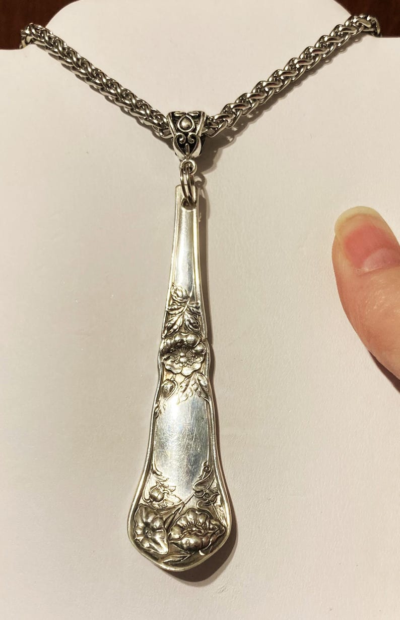 May include: A silver necklace with a long, ornate pendant made from a vintage silverware piece. The pendant features a floral design and is attached to a silver chain.