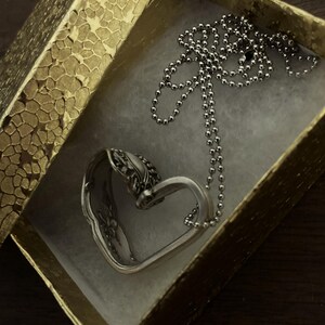 May include: A silver heart-shaped pendant necklace with a delicate chain. The necklace is in a gold gift box with a textured pattern.