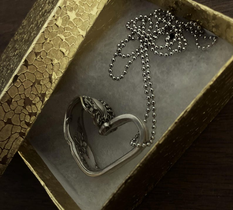 May include: A silver heart-shaped pendant necklace with a delicate chain. The necklace is in a gold gift box with a textured pattern.