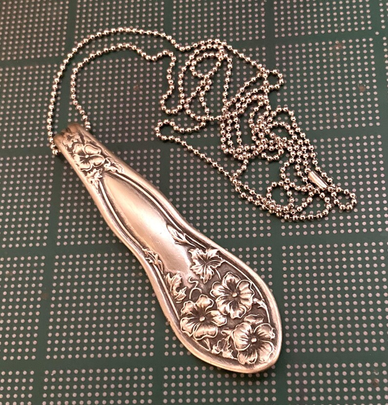 May include: A silver spoon necklace with a floral design. The spoon is attached to a silver ball chain.