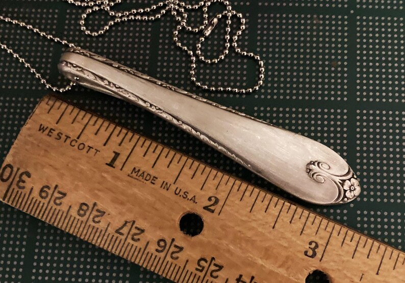 Puede incluir: Un collar de cuchara de plata con un dise&ntilde;o floral decorativo en el extremo. La cuchara mide aproximadamente 5 cm de largo y est&aacute; en una cadena de plata.