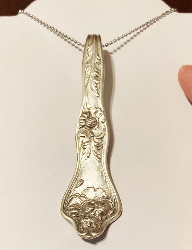 May include: A silver necklace with a pendant made from a vintage spoon. The spoon is engraved with a floral design and has a delicate chain.