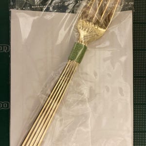 May include: A gold-colored fork with a green handle. The fork is wrapped in plastic and has a label that says "Resalable Cover Handle The Fork To Open".
