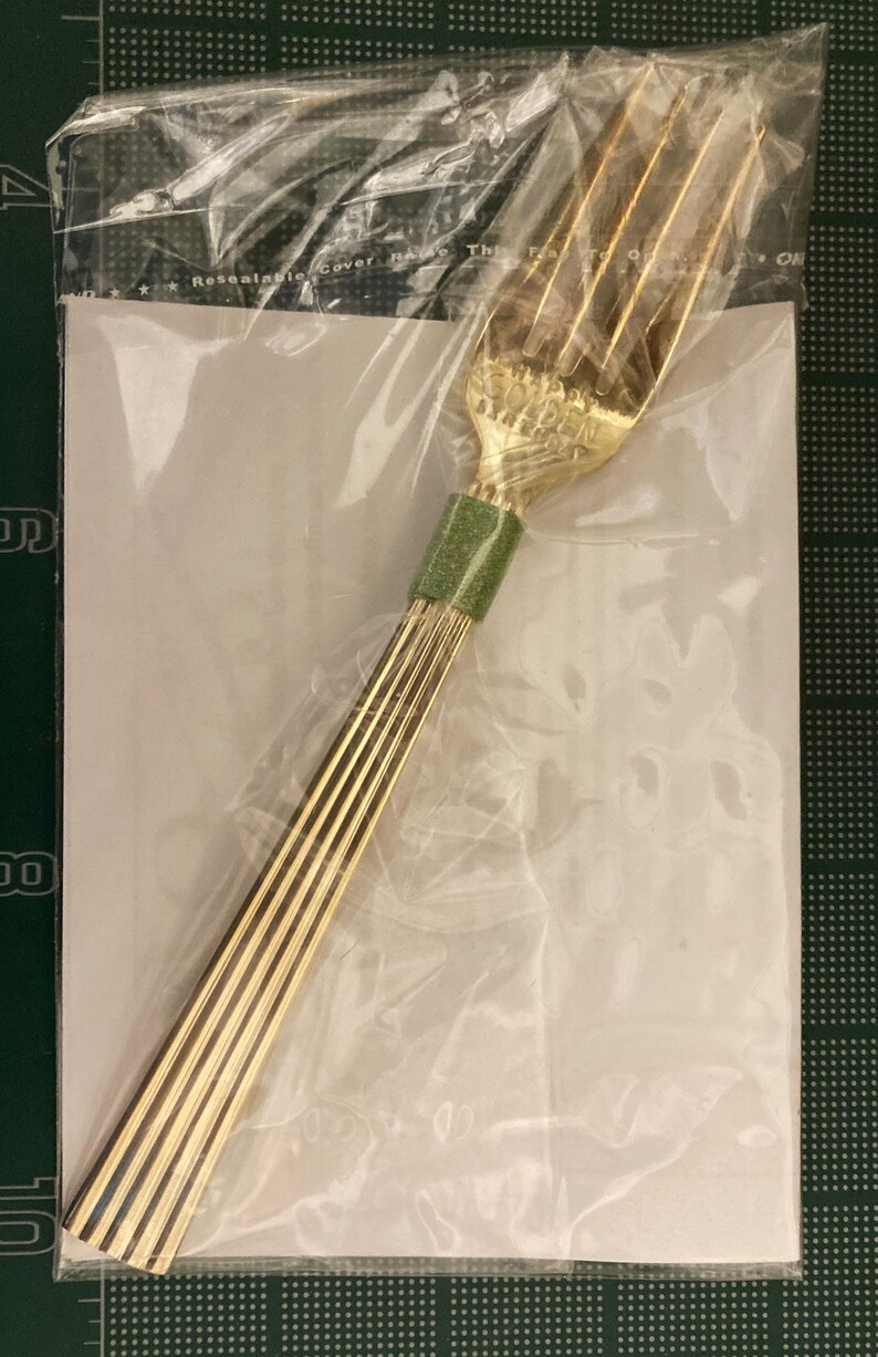May include: A gold-colored fork with a green handle. The fork is wrapped in plastic and has a label that says "Resalable Cover Handle The Fork To Open".