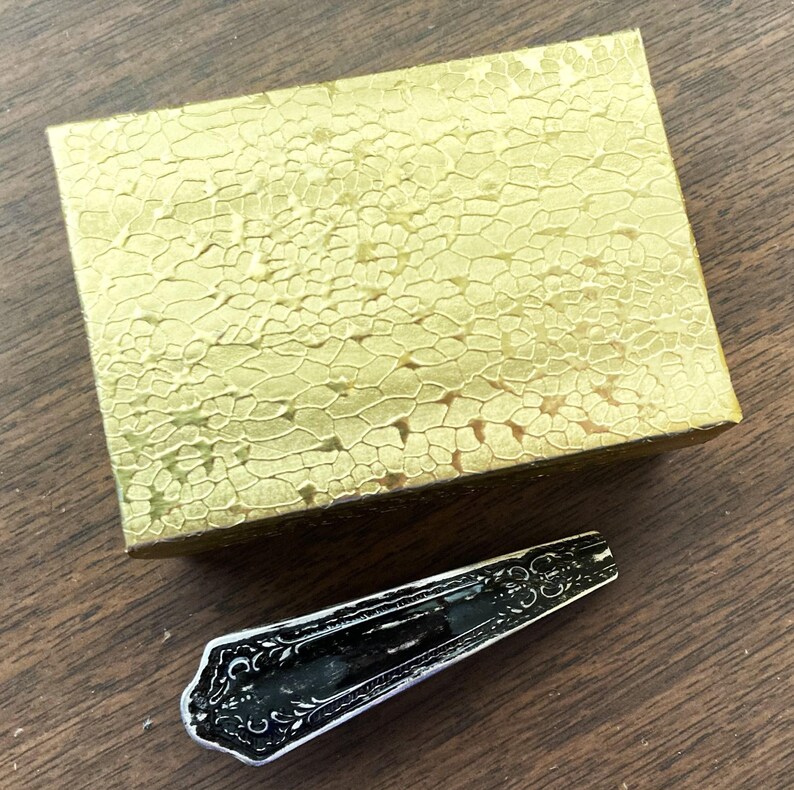 May include: A gold box with a textured surface and a black metal spoon with an ornate design.