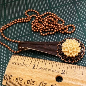 May include: A copper chain necklace with a brown wooden pendant and a cream-coloured flower charm. The flower charm is surrounded by a copper filigree design. The necklace is measured against a wooden ruler.