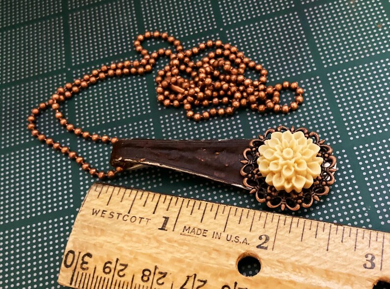May include: A copper chain necklace with a brown wooden pendant and a cream-coloured flower charm. The flower charm is surrounded by a copper filigree design. The necklace is measured against a wooden ruler.