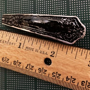 May include: A silver-toned, ornate, decorative spoon with a detailed design. The spoon is measured against a wooden ruler, showing it is approximately 1 inch long.
