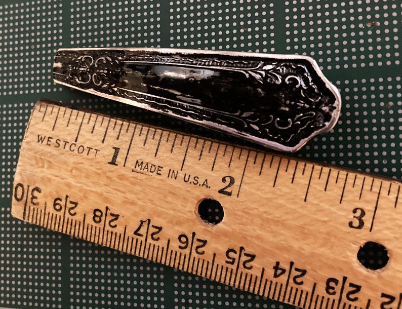 May include: A silver-toned, ornate, decorative spoon with a detailed design. The spoon is measured against a wooden ruler, showing it is approximately 1 inch long.