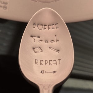 May include: A copper-colored spoon with the words "Coffee, Teach, Repeat" engraved on it. There is a small coffee cup, an open book, a pencil, and an arrow with a leaf on the spoon.