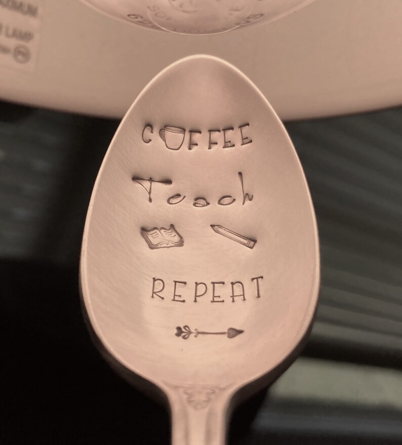 May include: A copper-colored spoon with the words "Coffee, Teach, Repeat" engraved on it. There is a small coffee cup, an open book, a pencil, and an arrow with a leaf on the spoon.