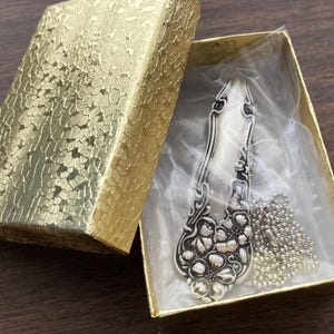May include: A silver spoon necklace with a floral design, packaged in a gold box with a clear plastic insert. The necklace is on a silver chain.