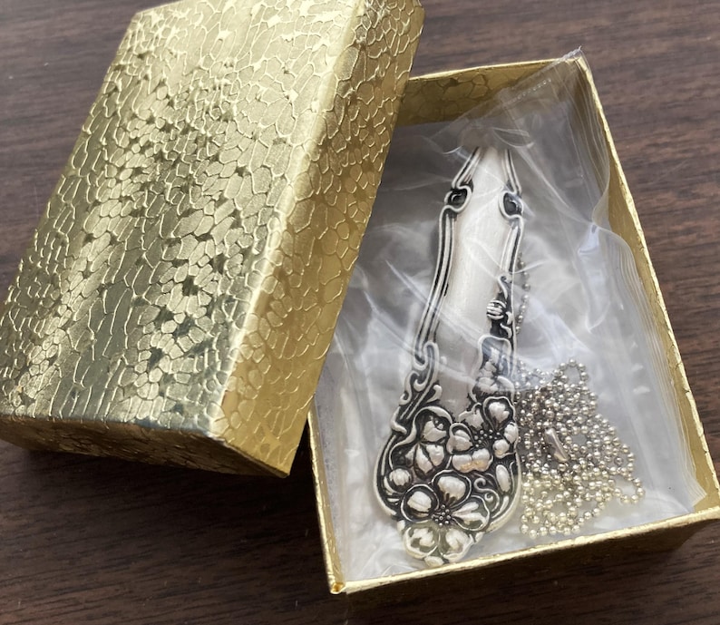 May include: A silver spoon necklace with a floral design, packaged in a gold box with a clear plastic insert. The necklace is on a silver chain.
