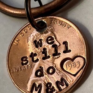 May include: A copper penny keychain with the words "We still do M&M" and a heart with the year 1983 engraved on it. The penny is attached to a copper key ring.